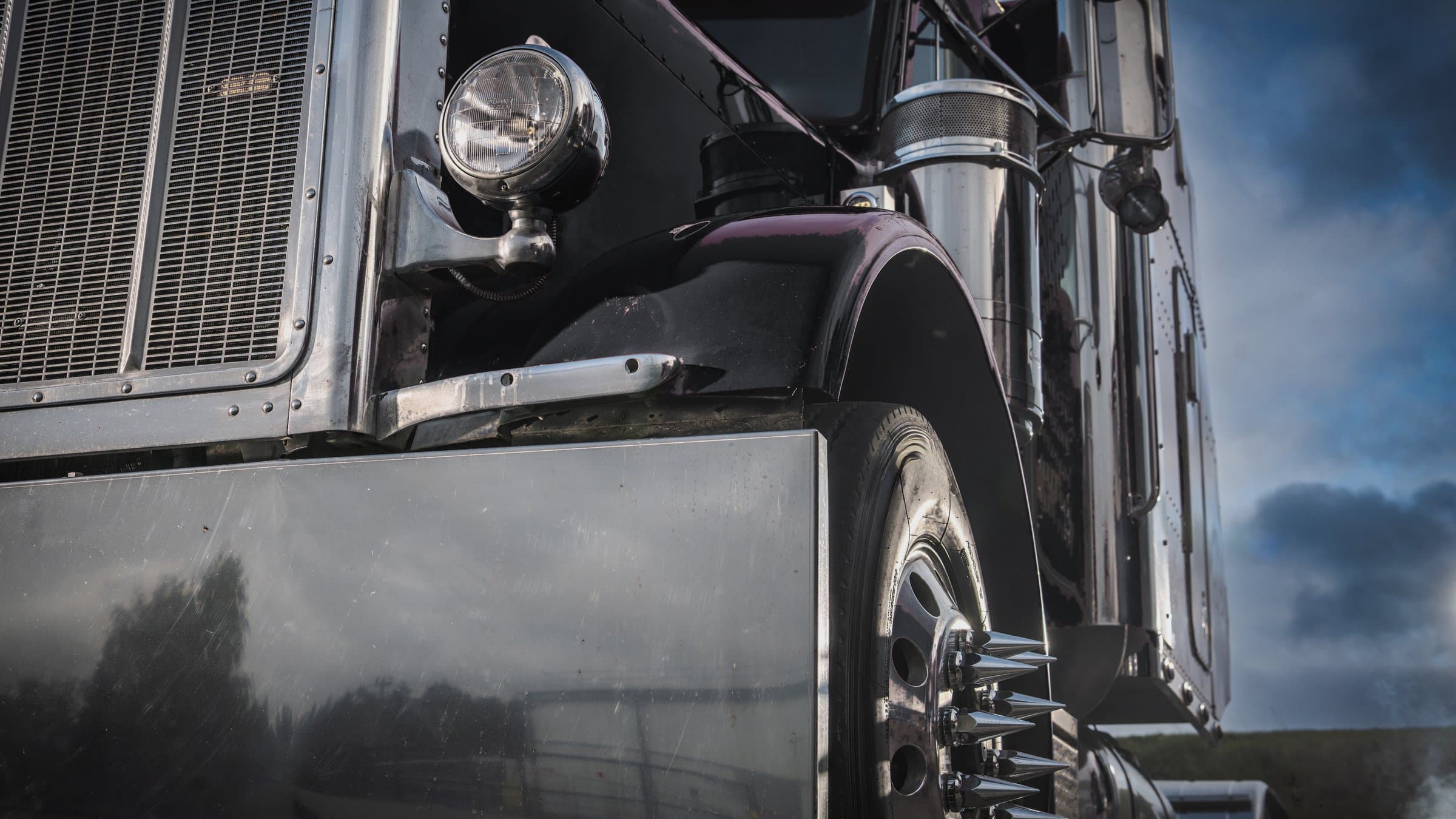 The front of a black classic semi truck under a storm sky, close on the headlight, fender and chrome stacks